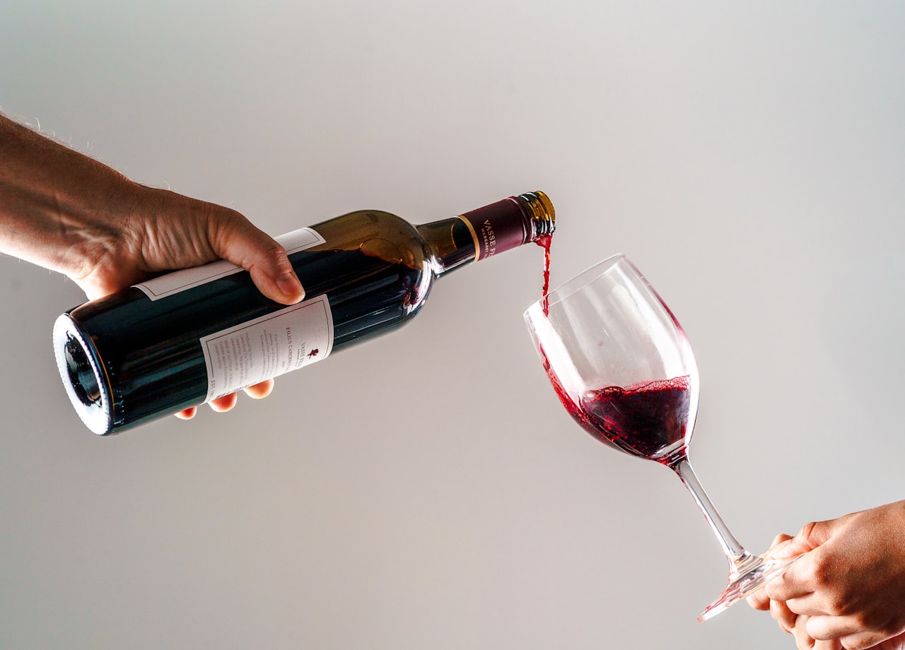 A host pouring red wine into a guest's glass at a holiday dinner table set with multiple open bottles and food